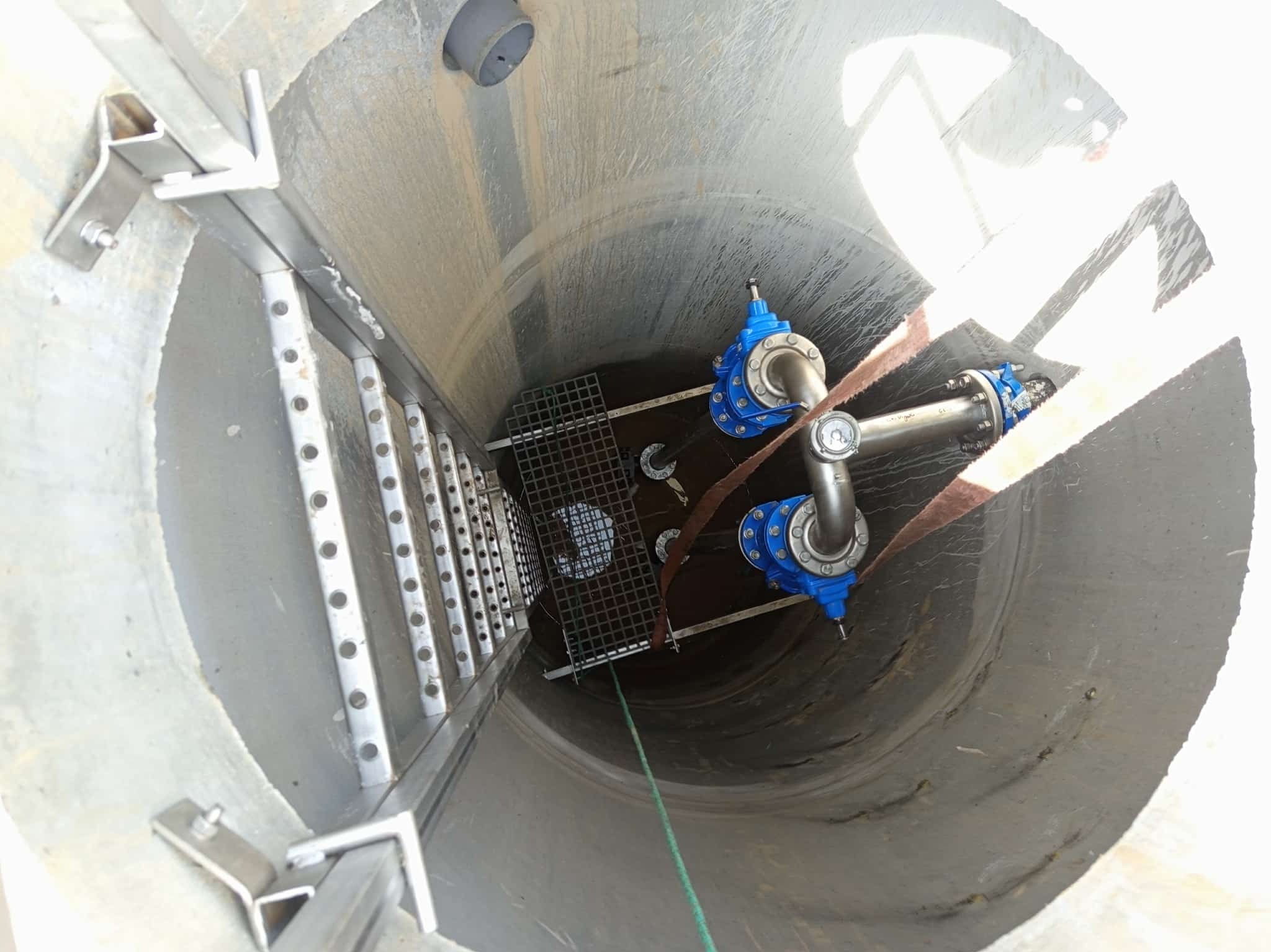 Wnętrze betonowego zbiornika z zainstalowaną drabinką i rurami z niebieskimi zaworami. Widok z góry, oświetlenie naturalne.