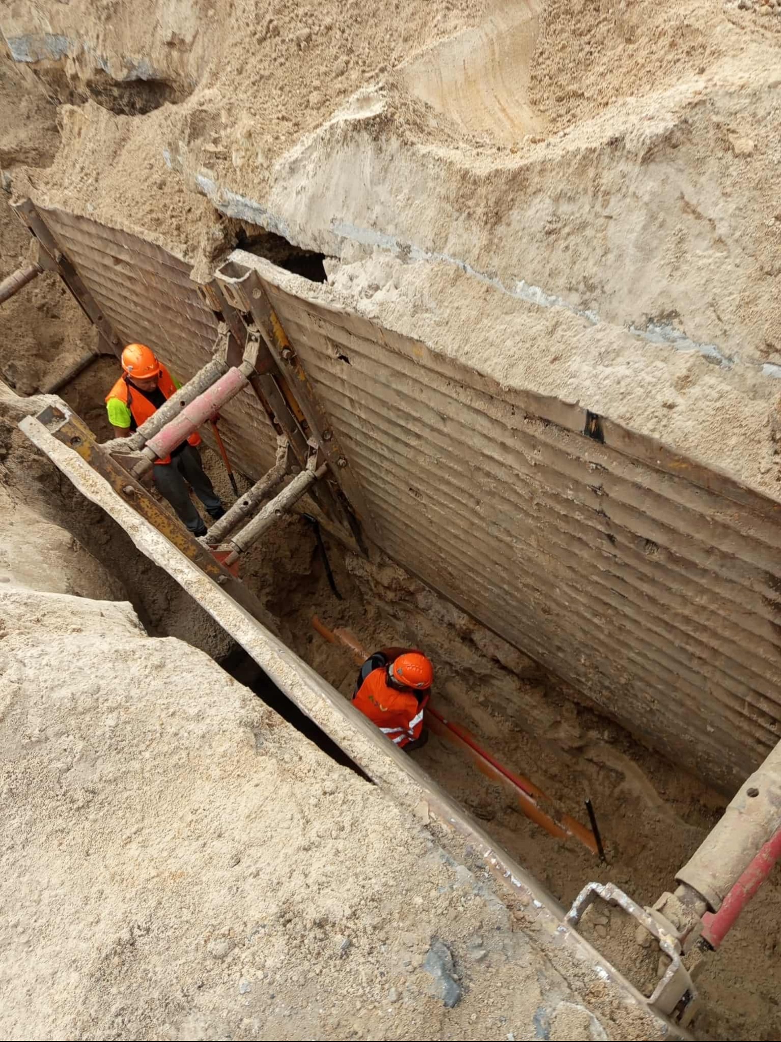 Głęboki wykop zabezpieczony szalunkiem stalowym. Dwóch robotników w kaskach i kamizelkach pracuje wewnątrz wykopu. Widok z góry na plac budowy.