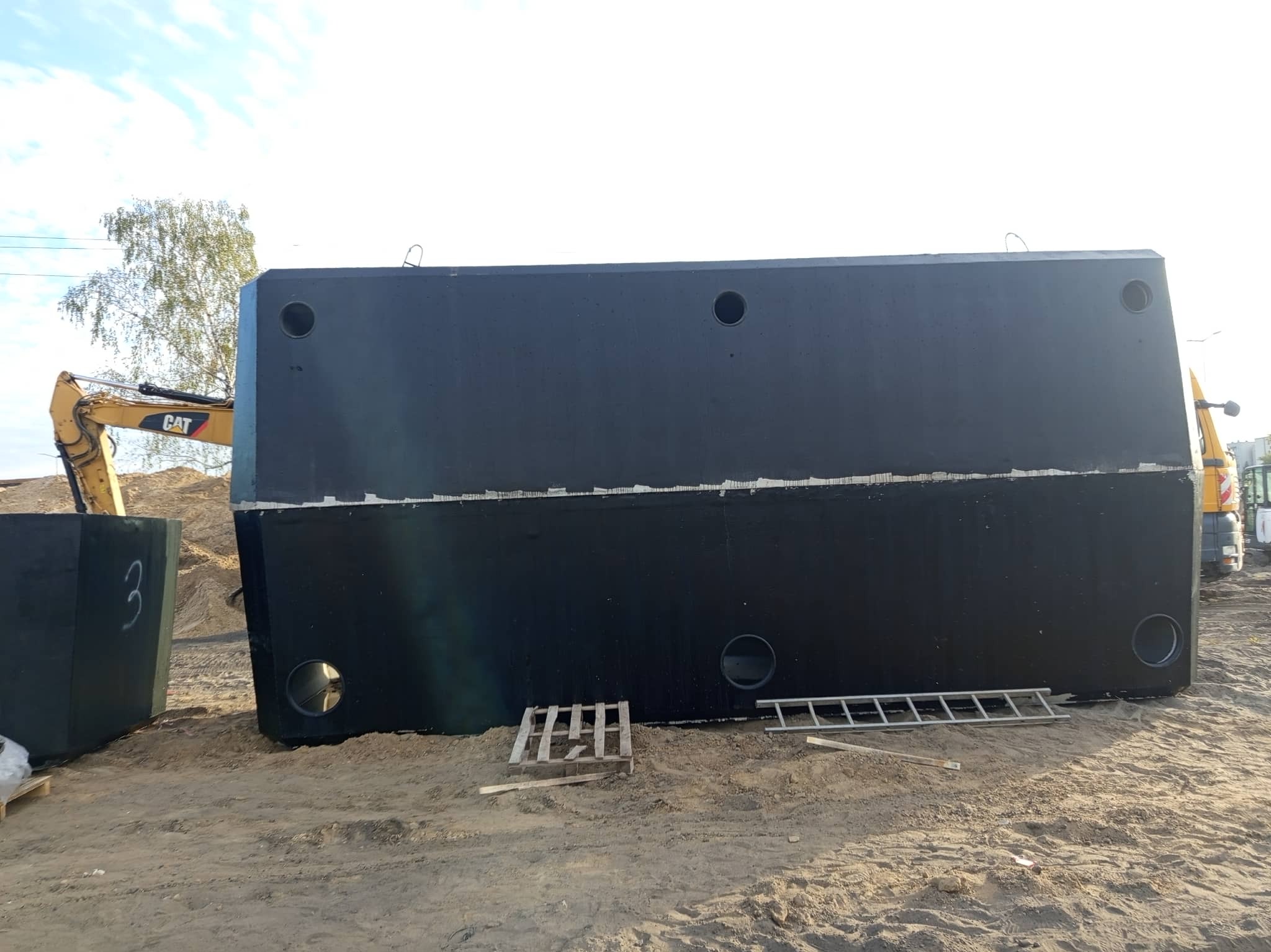 Dwa duże, czarne, betonowe zbiorniki z otworami, leżące na piasku. W tle widoczny żółty dźwig i drzewo. Na jednym ze zbiorników widnieje biała cyfra '3'.