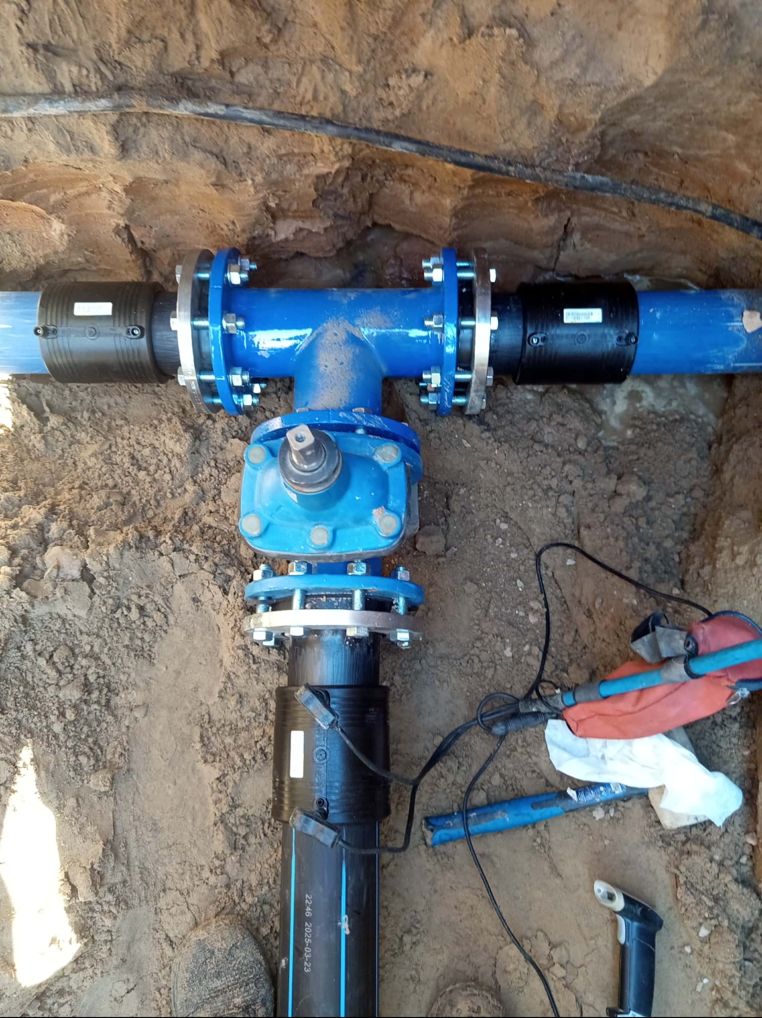 Niebieski trójnik z zaworem i złączkami kołnierzowymi, zainstalowany w wykopie ziemnym. Widoczne narzędzia i fragment stopy. Przyłącze wod-kan w trakcie realizacji.
