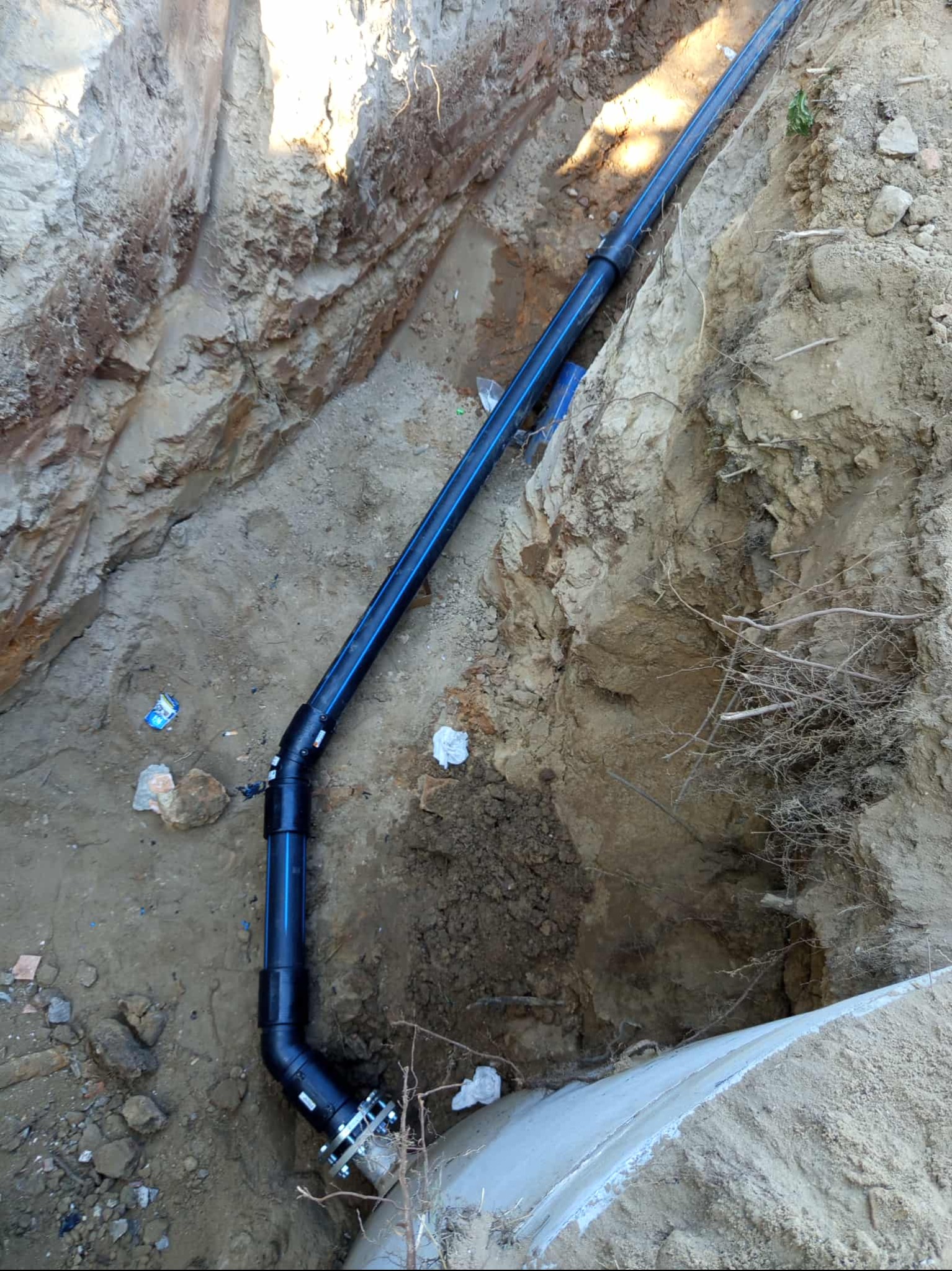 Niebieska rura kanalizacyjna ułożona w wykopie ziemnym, z widocznymi złączkami i elementami mocującymi. Wykop o nierównych ścianach, ziemia piaszczysta.