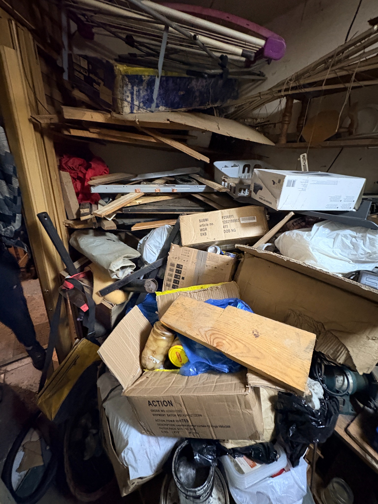 Zagracone pomieszczenie z kartonami, deskami i różnymi przedmiotami. Widoczny bałagan, wskazujący na potrzebę uprzątnięcia i wywozu zbędnych rzeczy.