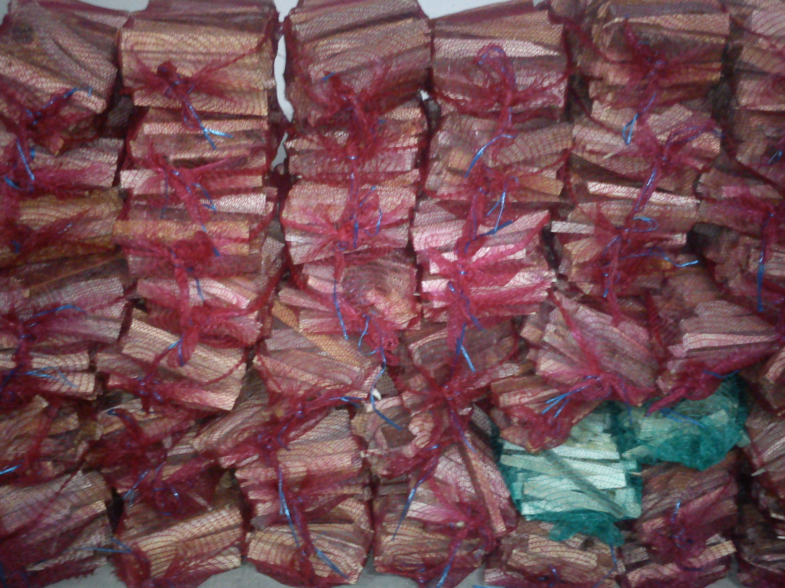 Stos worków z pociętym drewnem na rozpałkę, zapakowanych w siatki w kolorze bordowym i turkusowym, ułożonych jeden na drugim.