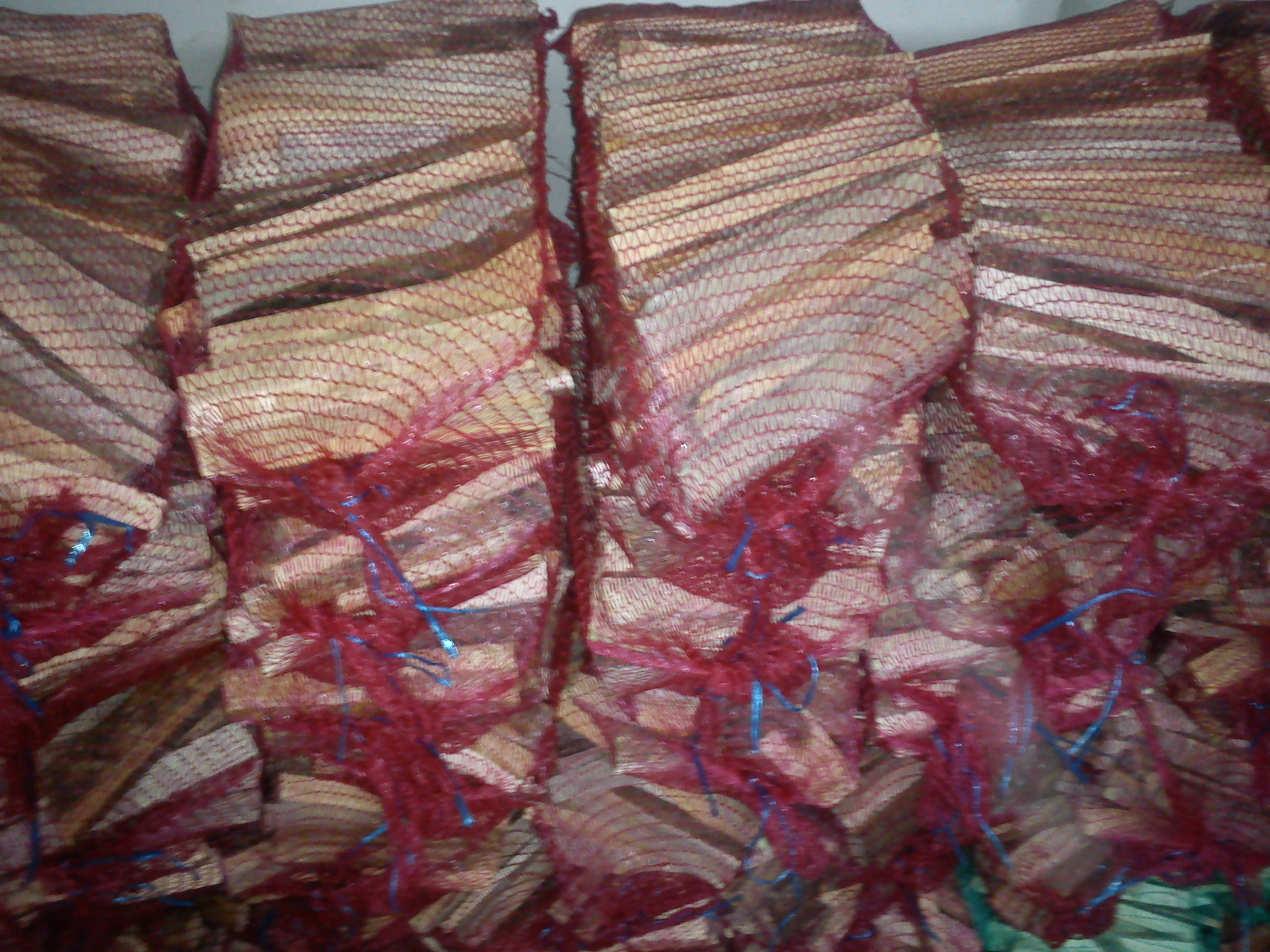 Ułożone worki z pociętym drewnem opałowym, zawiązane niebieskim sznurkiem w czerwonej siatce.