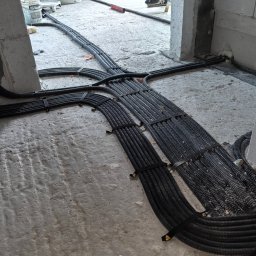 Robert Iwaniec - Rozłożone na surowym betonie, czarne, karbowane rury instalacji elektrycznej, spięte taśmami. Widoczne materiały budowlane i fragment osoby w tle.