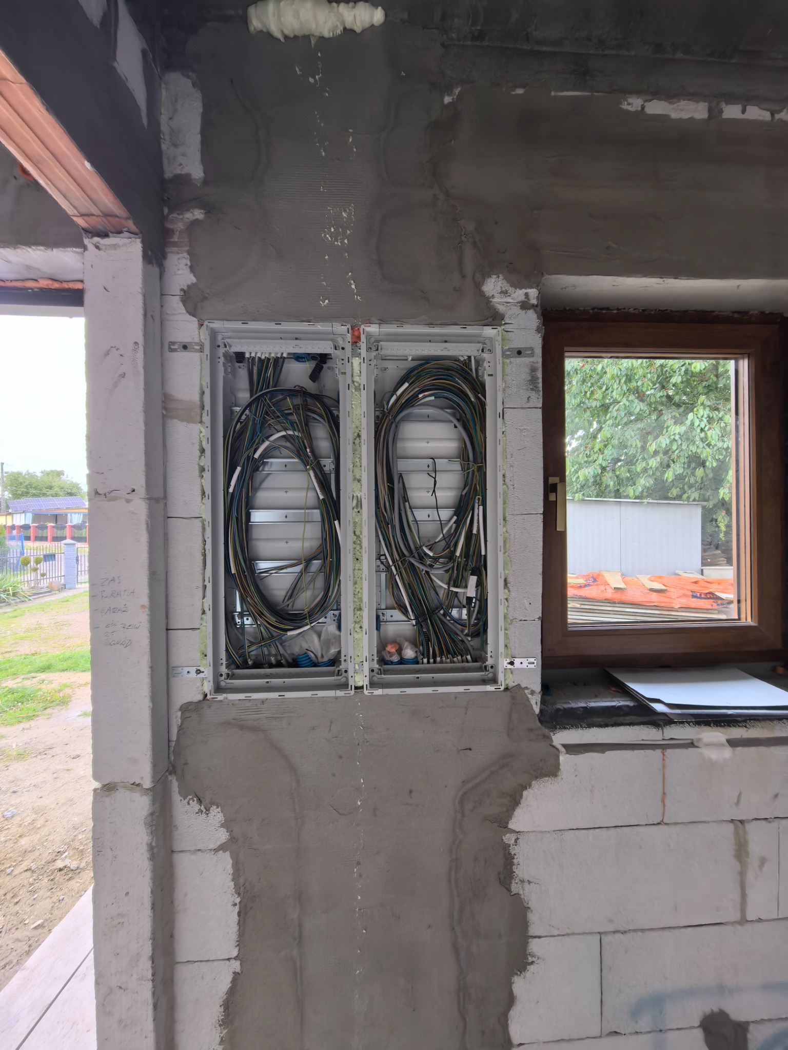 Dwa panele z kablami elektrycznymi w surowej ścianie z betonu komórkowego, widoczne okno z drewnianą ramą i elementy instalacji. Etap prac instalacyjnych.