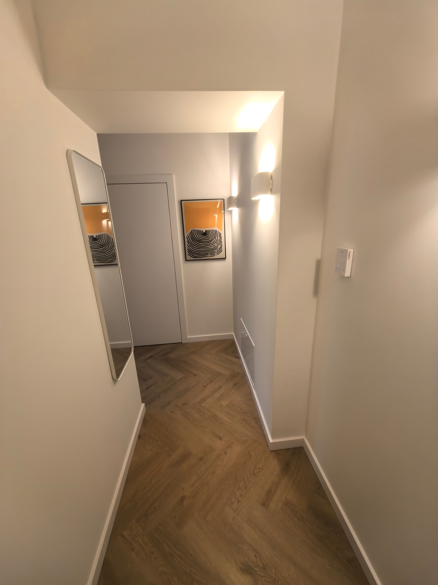 Korytarz z parkietem w jodełkę, oświetlony kinkietami. Na ścianie wisi lustro i obraz z motywem fal. Widoczny panel sterowania oświetleniem.