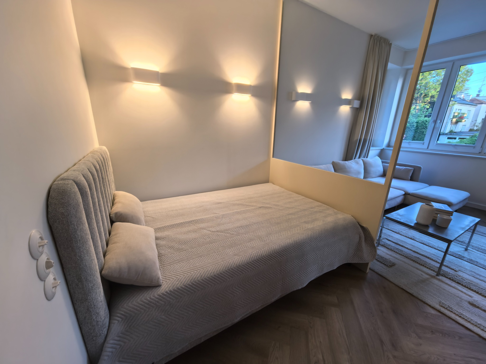 Nowoczesna sypialnia z podwójnym łóżkiem, oświetleniem ściennym i widokiem na salon. Na ścianie widoczne gniazdka elektryczne. Parkiet ułożony w jodełkę.