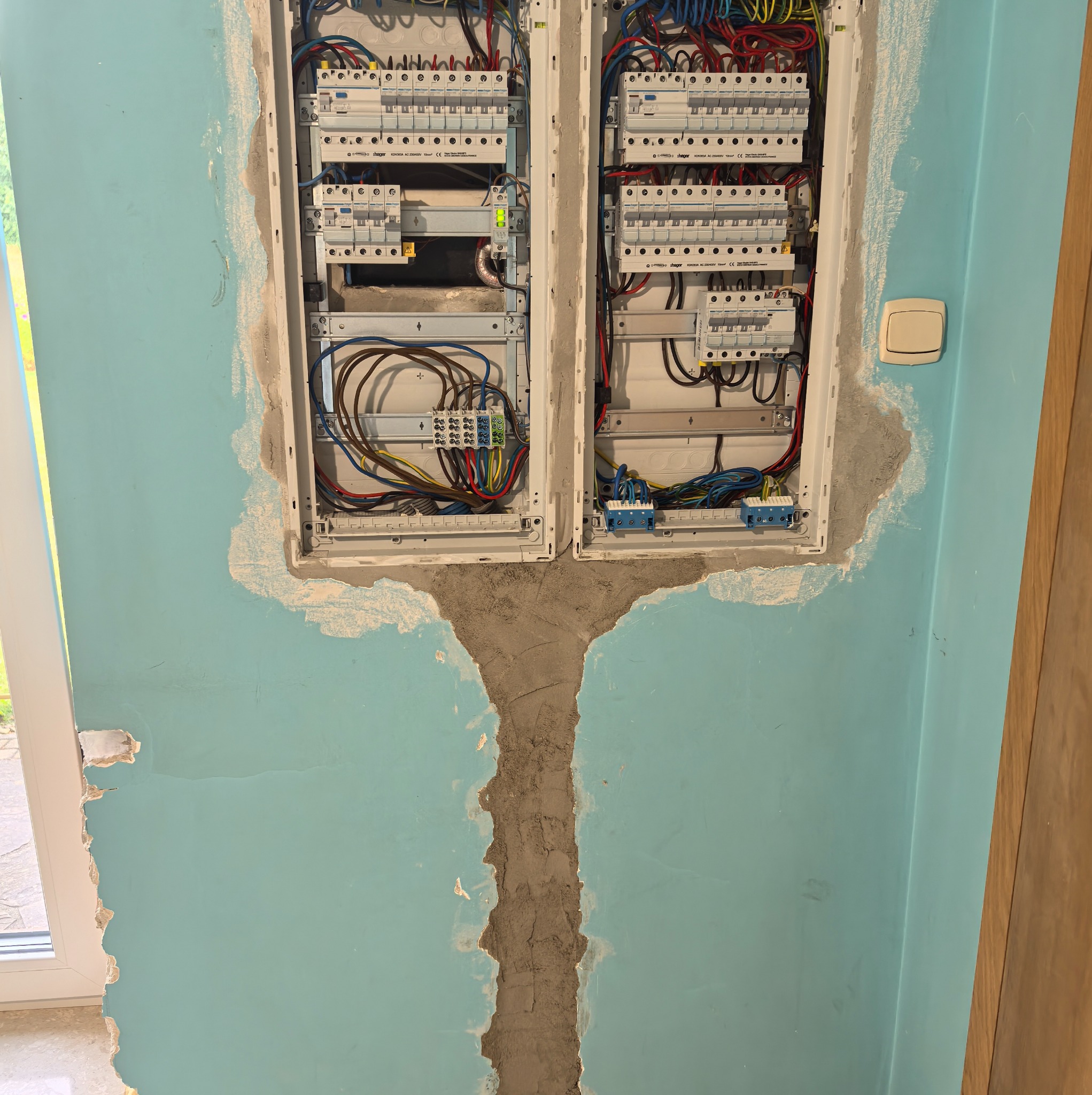 Dwa panele elektryczne w ścianie w trakcie montażu, widoczne kable i zabezpieczenia, ściana w kolorze błękitnym z widocznymi śladami po pracach montażowych.
