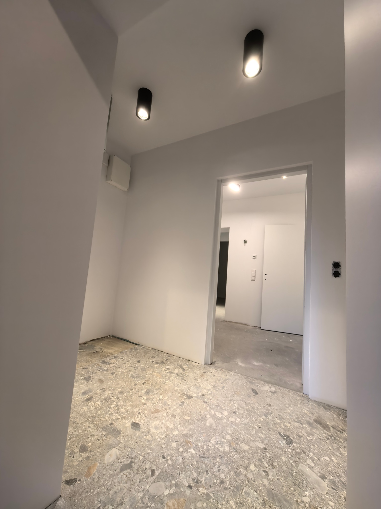 Wnętrze w trakcie remontu: szara podłoga z wzorem lastryko, białe ściany z zamontowanymi czarnymi lampami sufitowymi. Widoczne gniazdka i włączniki.