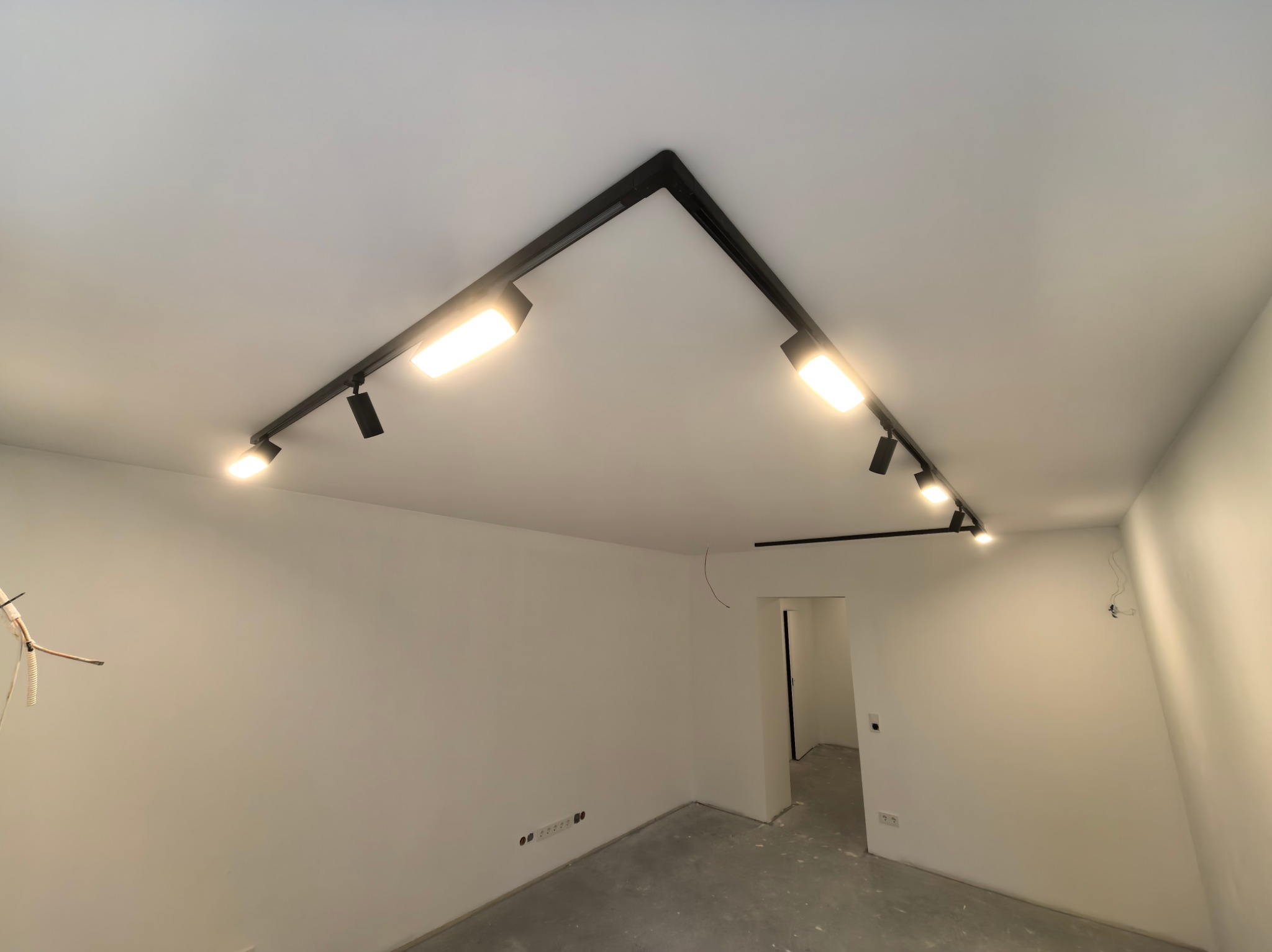 Czarna, szynowa instalacja oświetleniowa z reflektorami LED na białym suficie w surowym wnętrzu. Widoczne gniazdka elektryczne i niedokończone ściany.