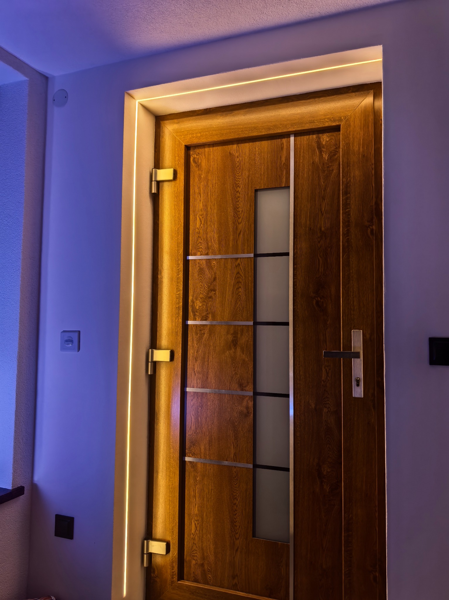 Drewniane drzwi wewnętrzne z oświetleniem LED wzdłuż futryny, tworzące ciepłą, nowoczesną atmosferę. Widoczne zawiasy i klamka.