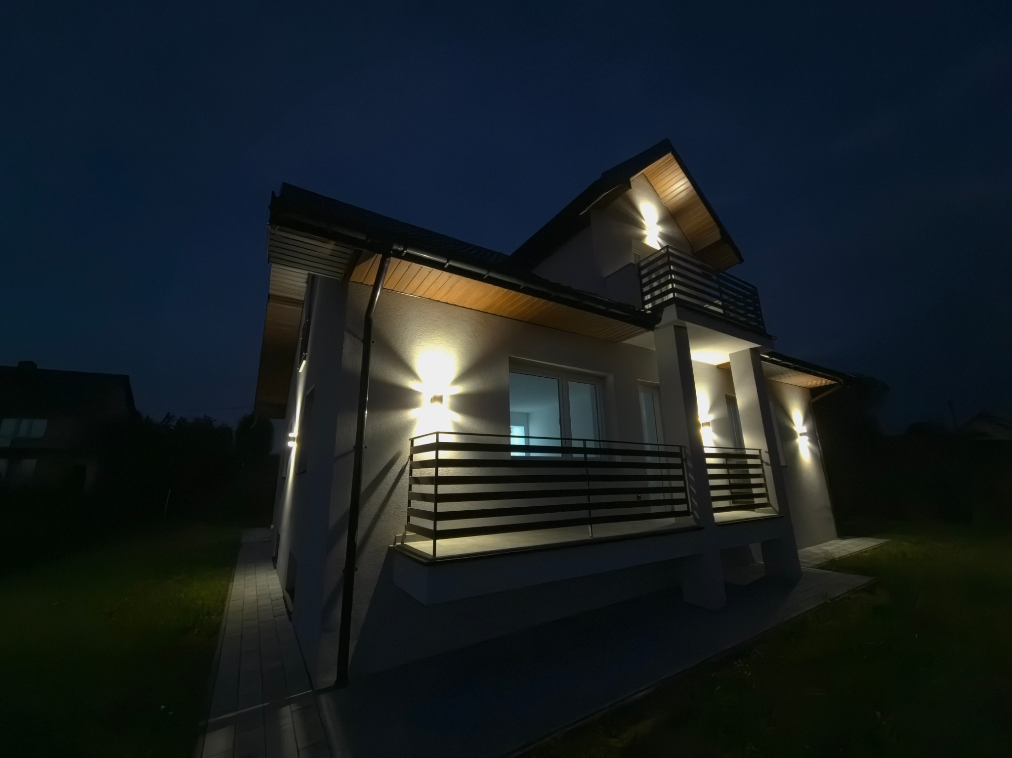 Nowoczesny dom z balkonami oświetlony zewnętrznymi lampami ściennymi, nocne ujęcie z dołu, widoczna elewacja i fragment chodnika.