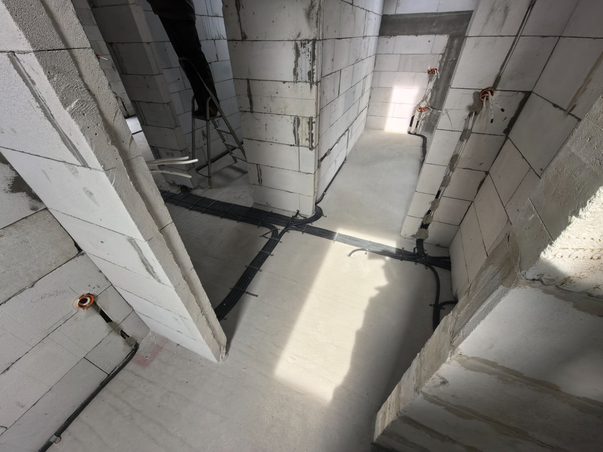 Surowy stan budynku z widoczną instalacją elektryczną w korytach kablowych na podłodze i wyprowadzonymi przewodami ze ścian. Widoczne bloczki betonowe i drabina.