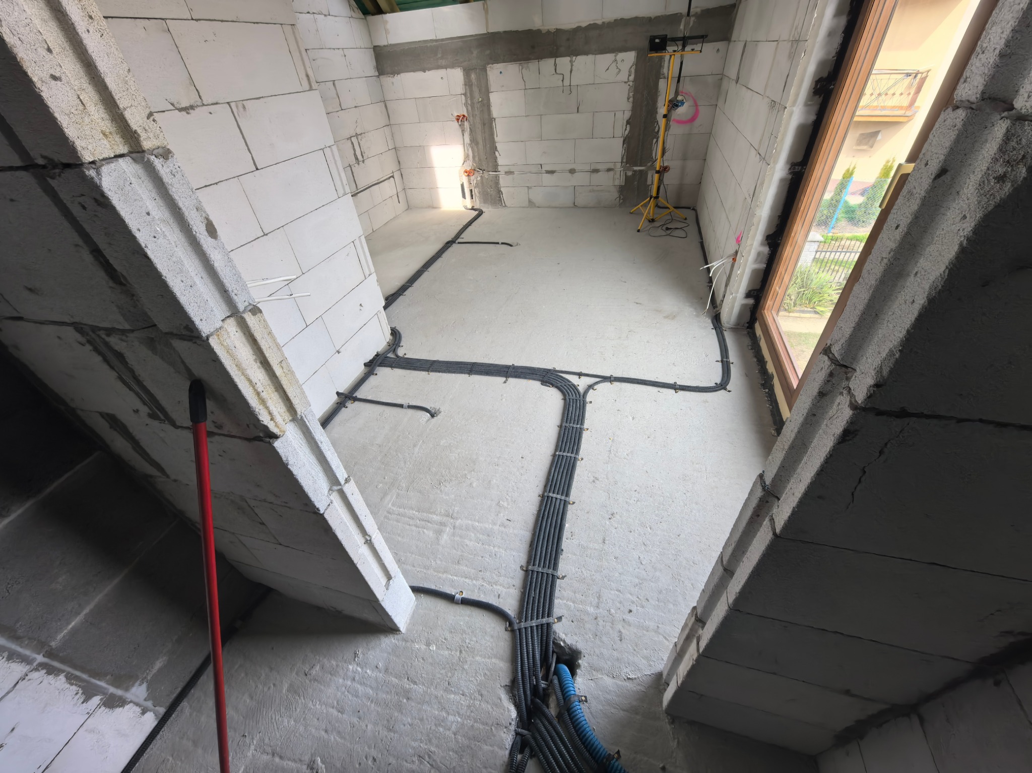 Wnętrze w trakcie prac elektrycznych: kable w peszlach ułożone na podłodze, ściany z betonu komórkowego, widoczne puszki instalacyjne i lampa robocza.