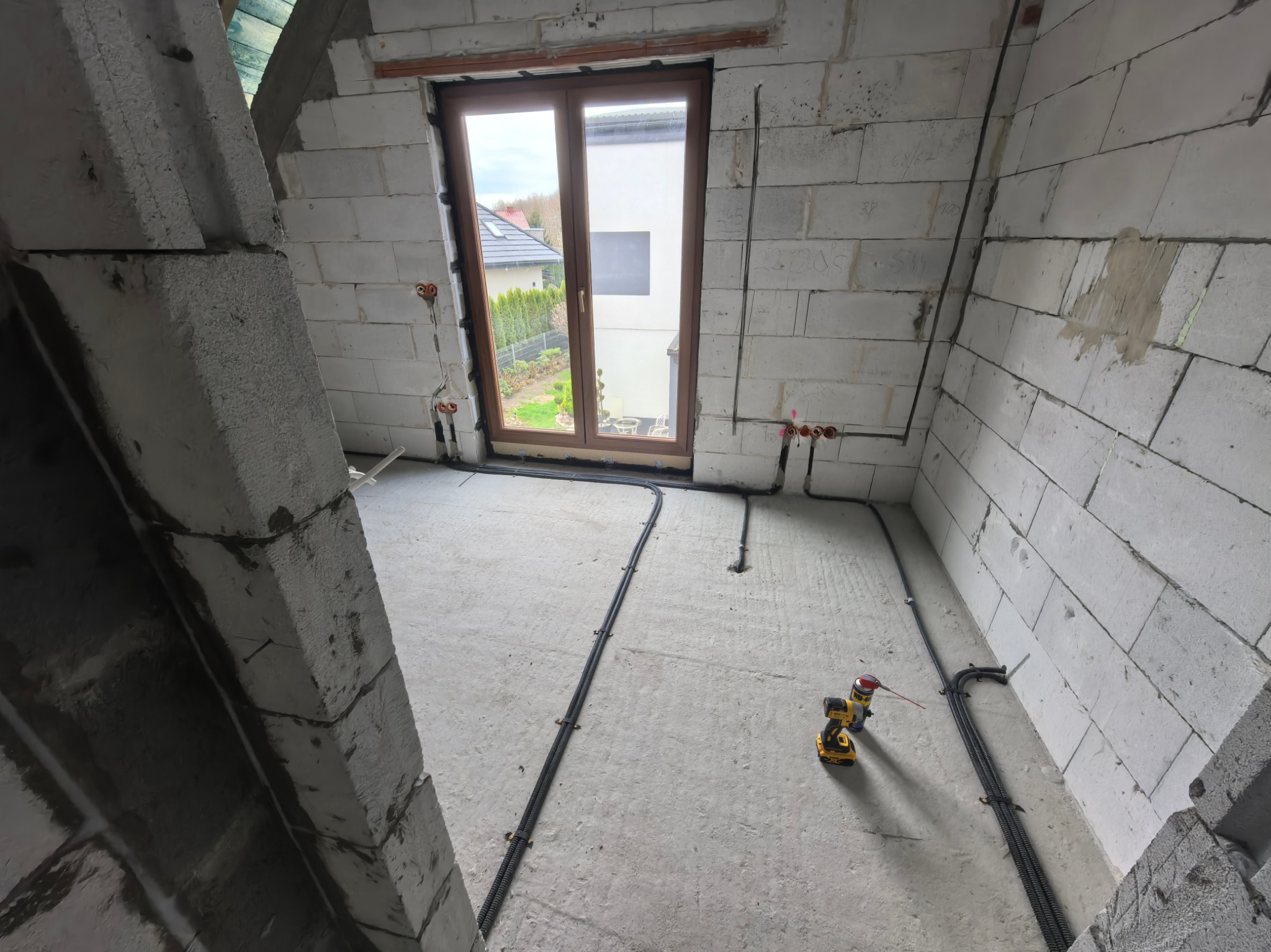 Wnętrze w trakcie instalacji elektrycznej: kable w peszlach ułożone na podłodze, puszki elektryczne na ścianach z betonu komórkowego, okno w tle. Widoczna wiertarko-wkrętarka.
