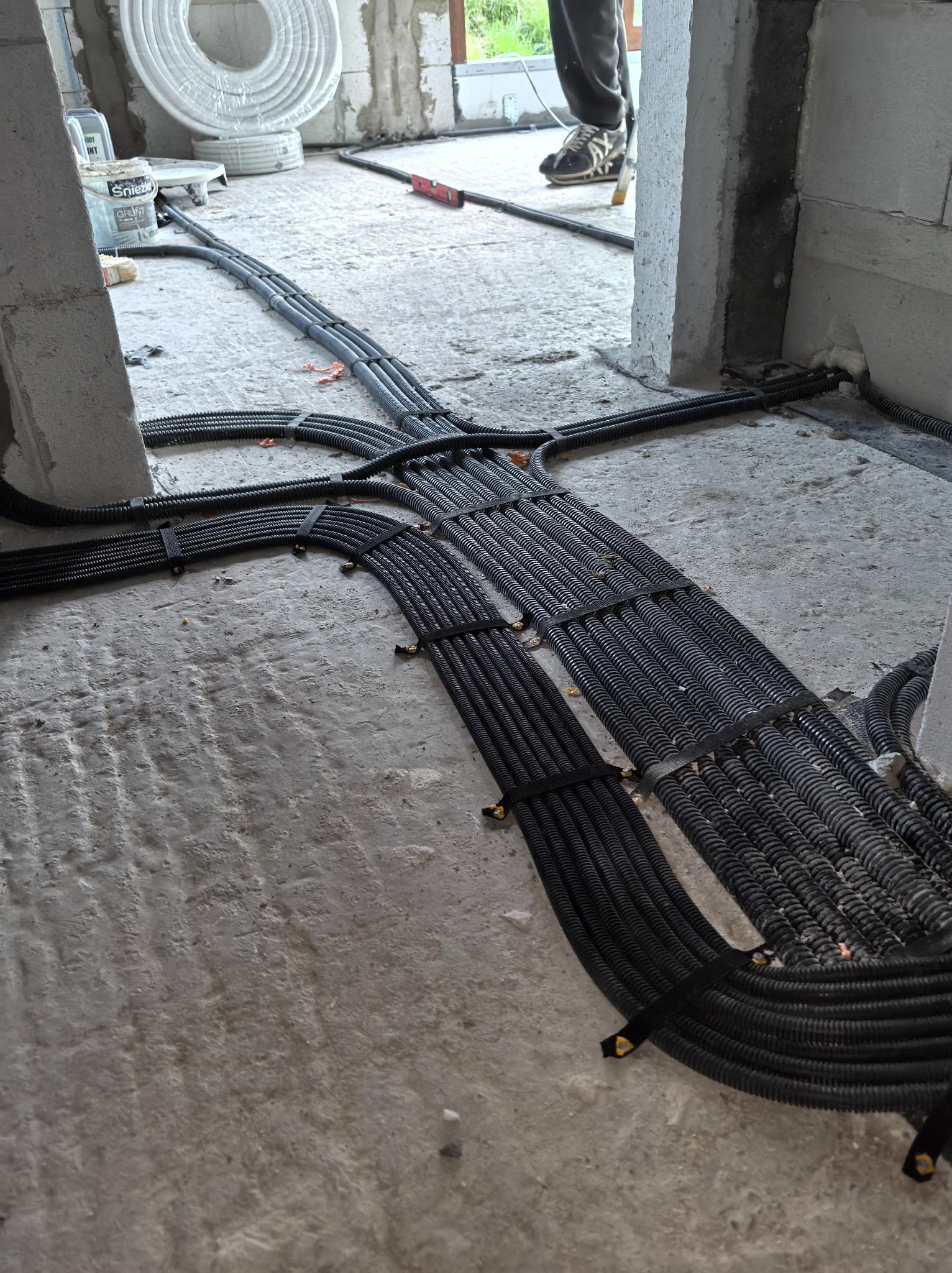Rozłożone na surowym betonie, czarne, karbowane rury instalacji elektrycznej, spięte taśmami. Widoczne materiały budowlane i fragment osoby w tle.