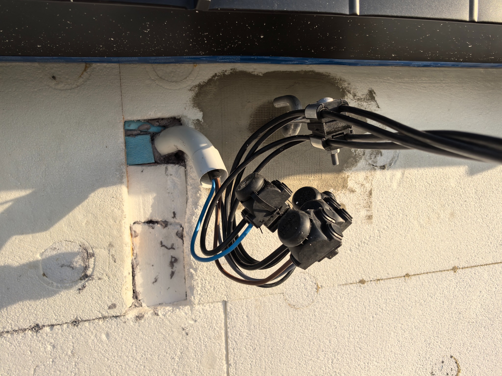 Podłączenie kabli elektrycznych do rury w ścianie ocieplonej styropianem. Widoczne przewody w kolorach niebieskim i brązowym. Elementy instalacji zabezpieczone uchwytami.