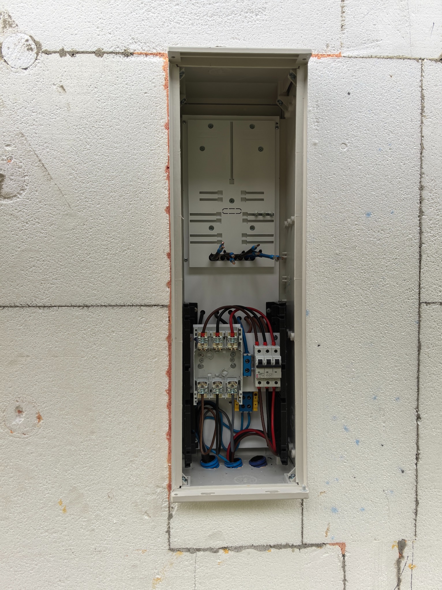 Rozdzielnica elektryczna wnękowa, zamontowana w ścianie z białego styropianu. Widoczne przewody, bezpieczniki i elementy montażowe. Ujęcie z bliska, oświetlenie naturalne.