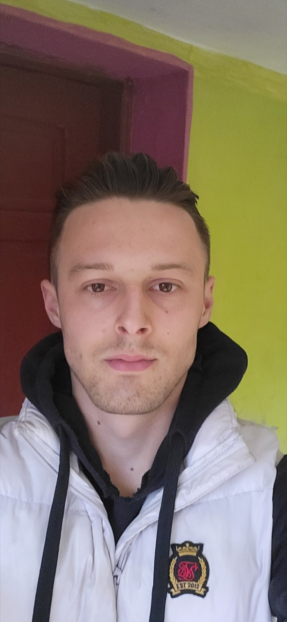 Portret młodego mężczyzny w białej kamizelce na tle ściany w dwóch kolorach: czerwonym i zielonym. Ujęcie od przodu, skupione na twarzy i górnej części ciała.