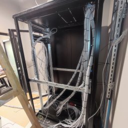 Pavel Skarynka - Metalowa szafa rack z uporządkowanymi kablami wewnątrz, zabezpieczonymi czarnymi opaskami. Widoczna drabina malarska i klimatyzator na górze szafy.