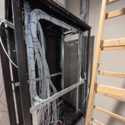 Pavel Skarynka - Rozbudowa szafy serwerowej z uporządkowanymi kablami w białych peszlach, widoczne drabinki kablowe i drabina drewniana. Etap prac w pomieszczeniu technicznym.