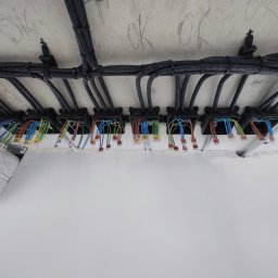 Pavel Skarynka - Rozprowadzenie kabli w czarnych peszlach, zamocowanych do sufitu, z widocznymi kolorowymi przewodami i złączkami w puszkach instalacyjnych. Widok z dołu.