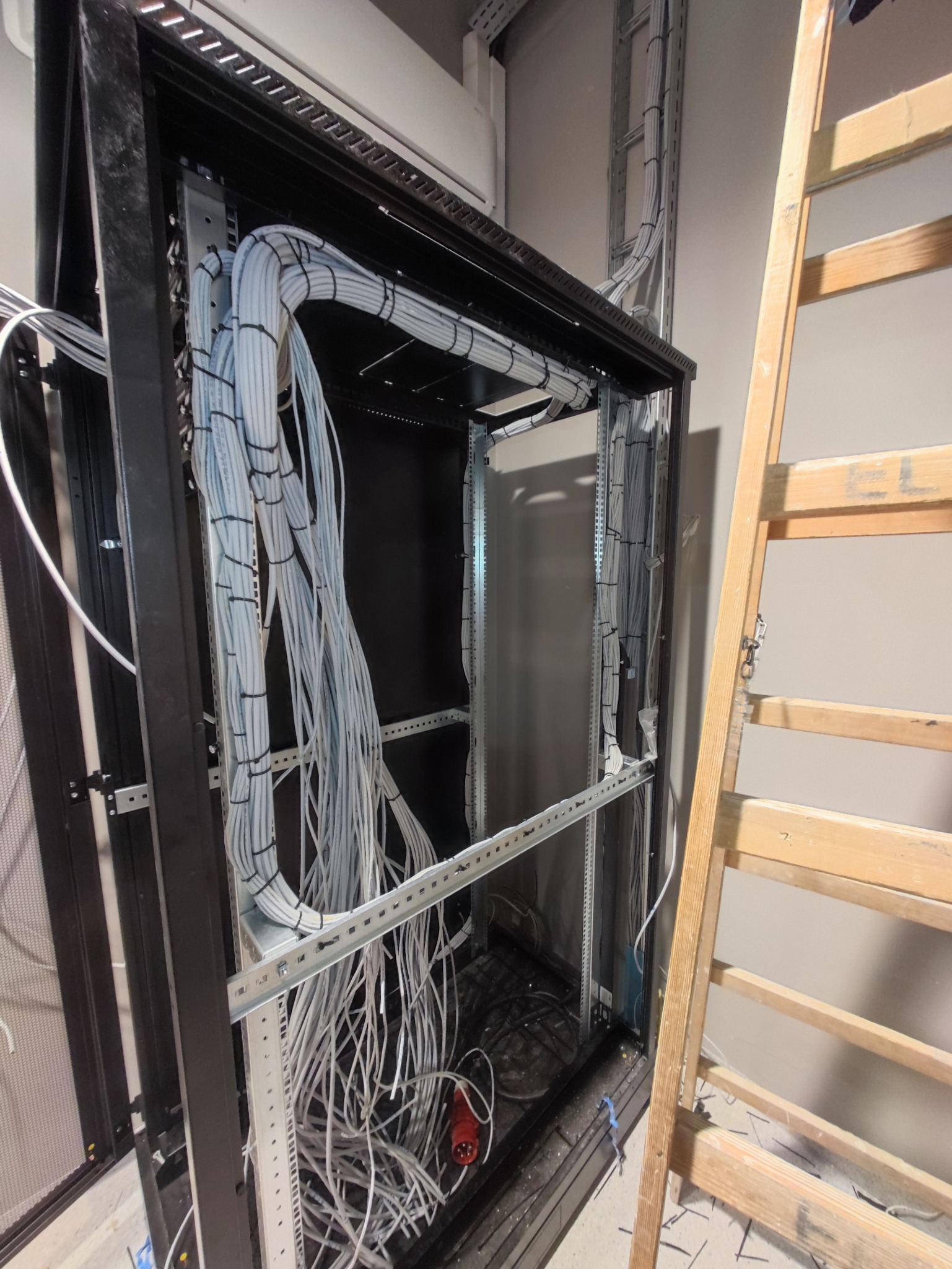 Rozbudowa szafy serwerowej z uporządkowanymi kablami w białych peszlach, widoczne drabinki kablowe i drabina drewniana. Etap prac w pomieszczeniu technicznym.