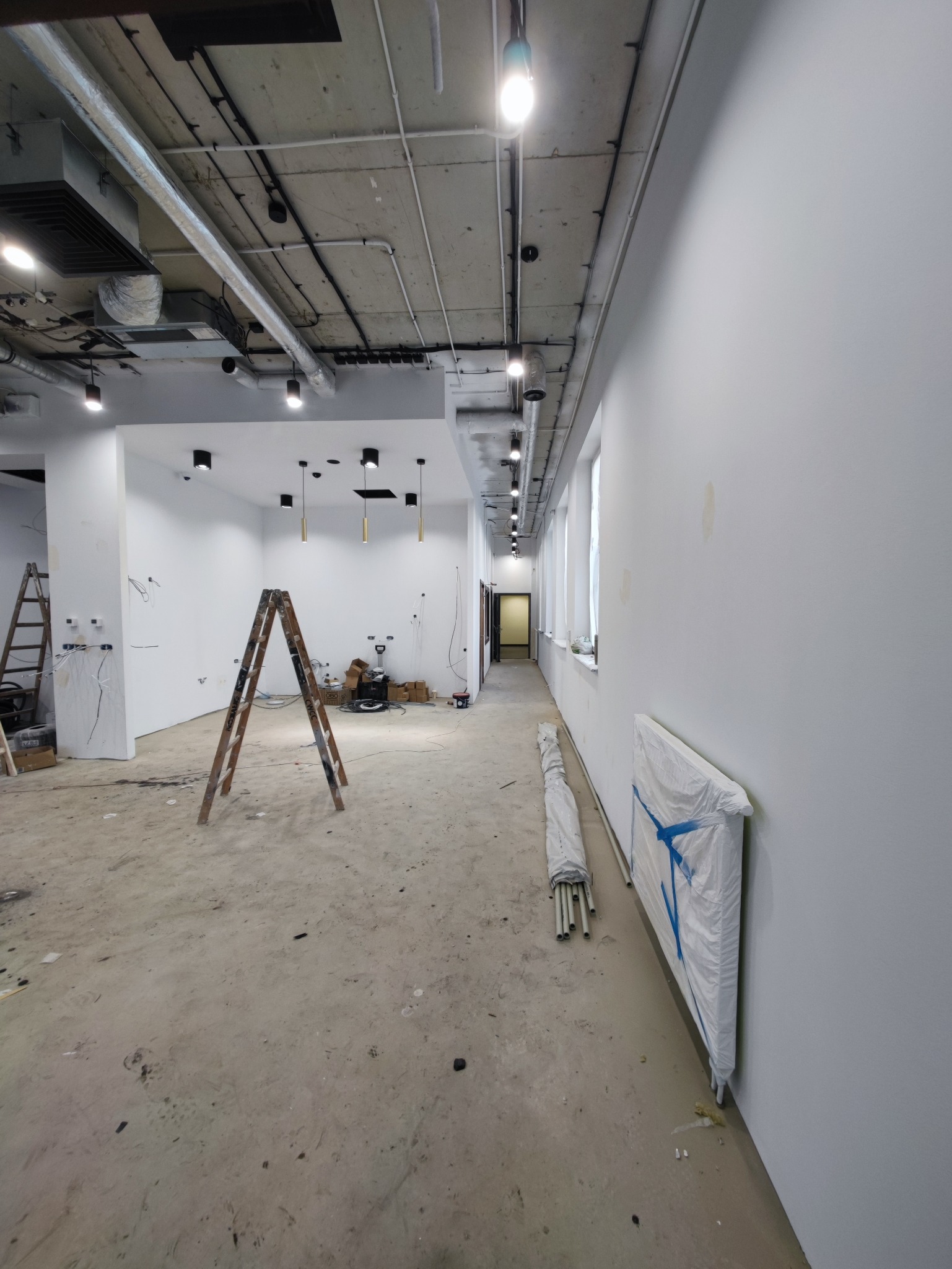 Wnętrze w trakcie prac wykończeniowych z widocznymi lampami sufitowymi, przewodami i drabinami. Stan surowy podłogi. Widok na korytarz z oknami.