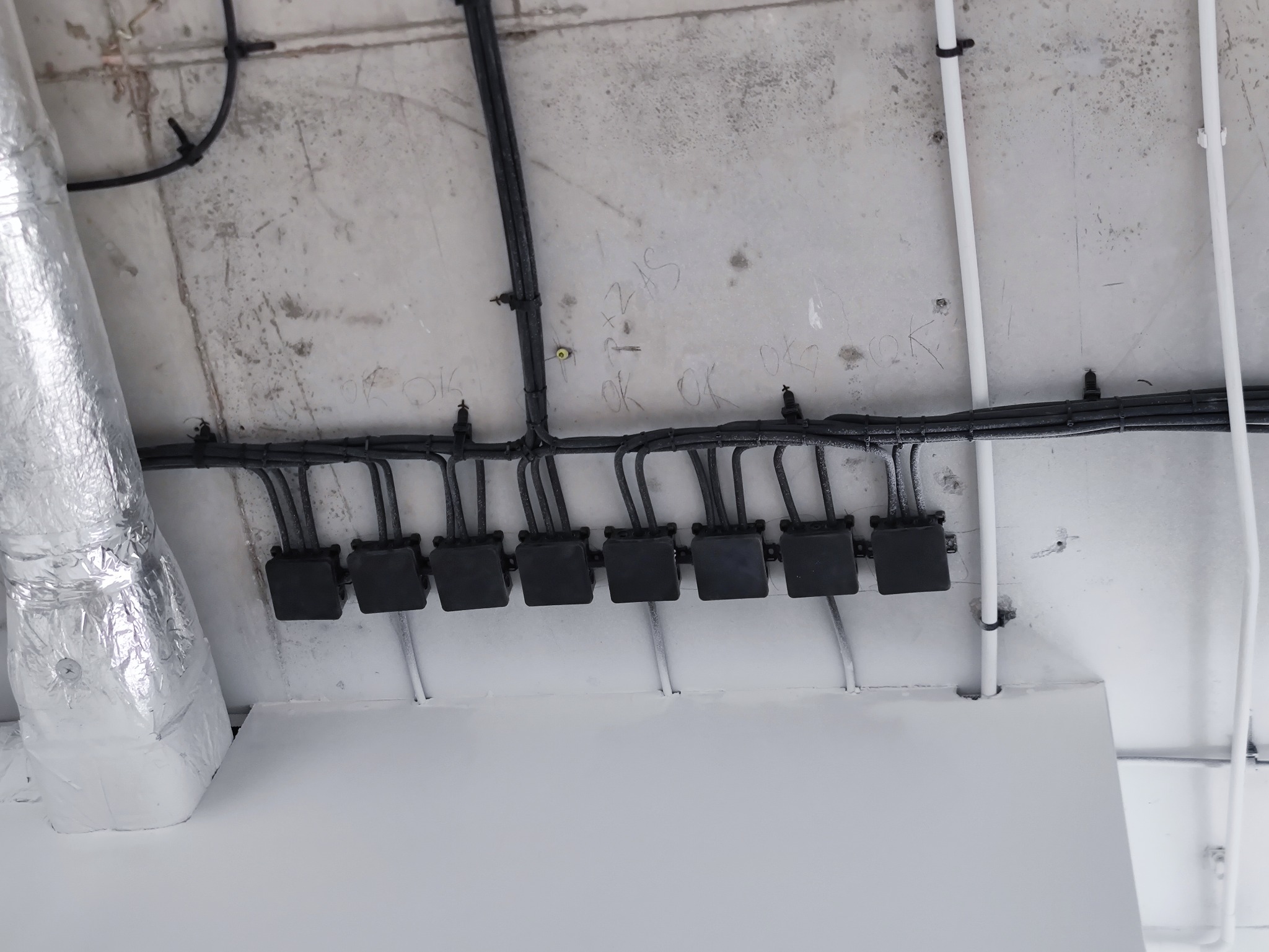 Szereg czarnych puszek instalacyjnych pod sufitem z widocznymi przewodami i rurą wentylacyjną w tle. Widoczne ślady po instalacjach na suficie.