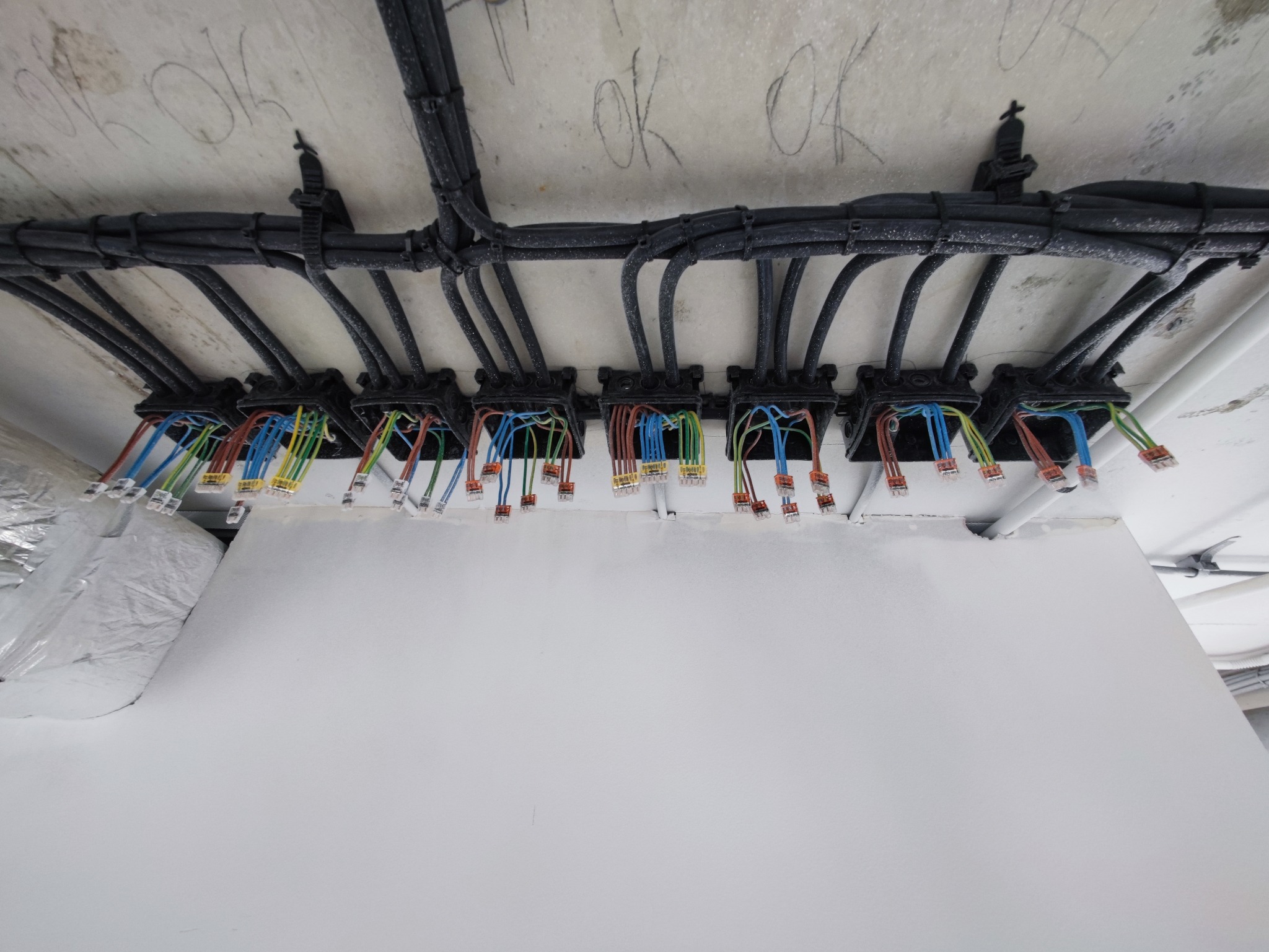 Rozprowadzenie kabli w czarnych peszlach, zamocowanych do sufitu, z widocznymi kolorowymi przewodami i złączkami w puszkach instalacyjnych. Widok z dołu.