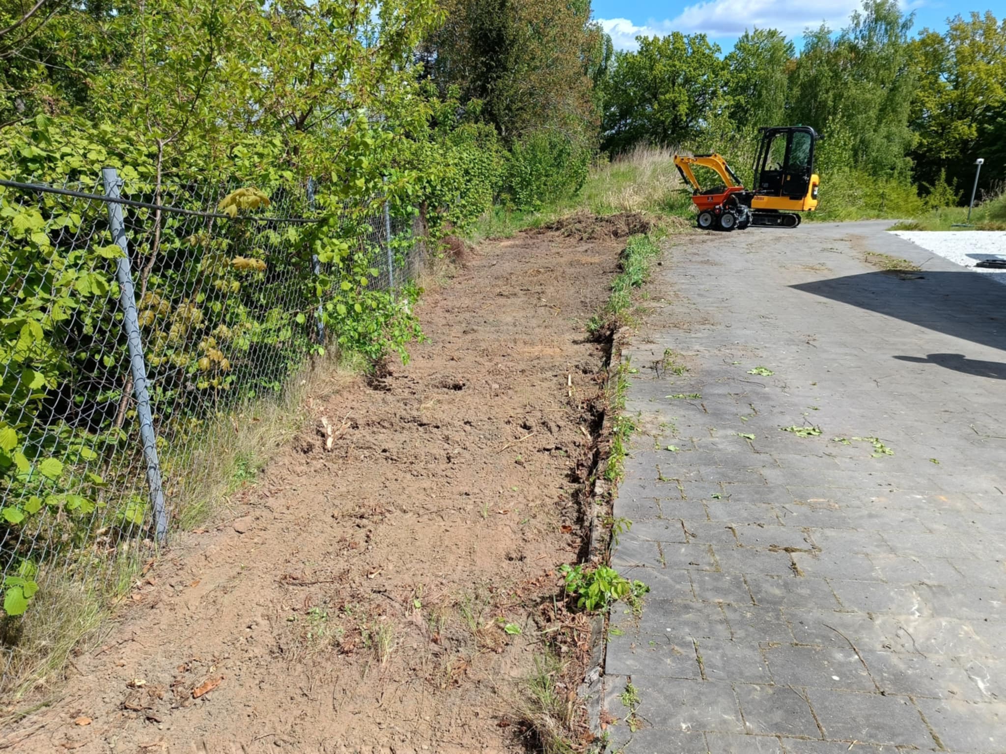 Teren przygotowany pod przyszłe prace ziemne, widoczny mały traktor, siatka ogrodzeniowa i fragment chodnika z kostki. Etap początkowy prac.