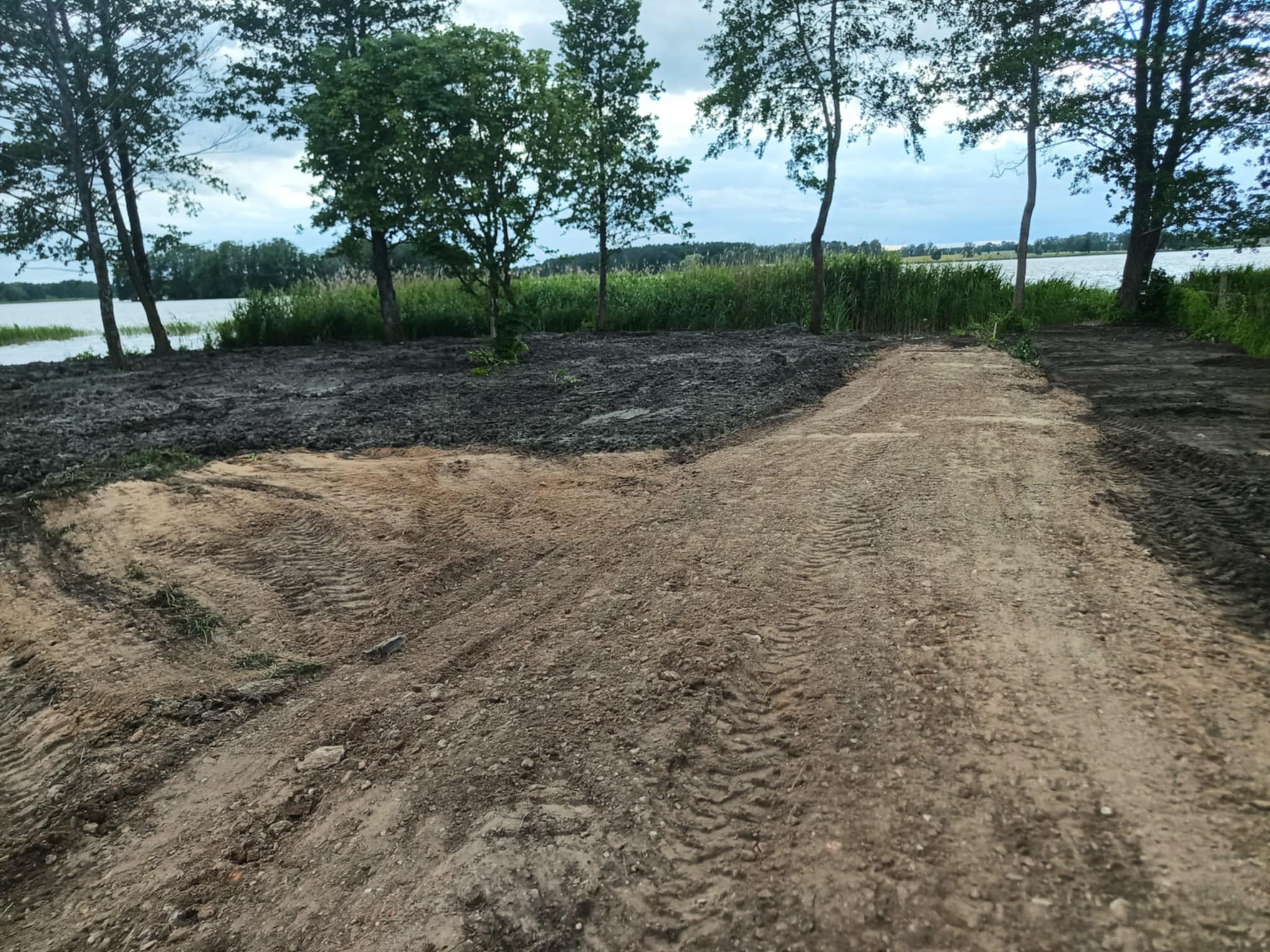 Przygotowany teren pod przyszły ogród z widocznymi śladami opon, częściowo z czarną ziemią, w tle jezioro i drzewa. Etap prac ziemnych przed założeniem ogrodu.
