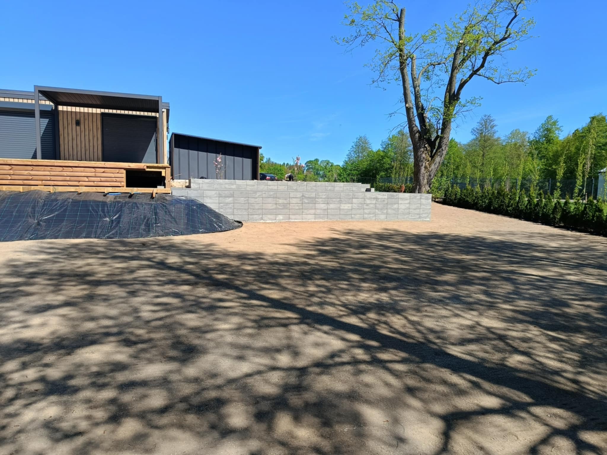 Przygotowanie terenu pod ogród: wyrównana ziemia, murki oporowe z szarej kostki, widoczne drzewa i elementy architektury ogrodowej w słoneczny dzień.