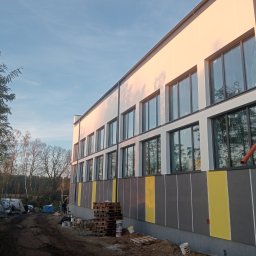 Usługi Remontowo-Wykończeniowe Damian Zaborowski - Nowoczesny budynek z oknami i elewacją w odcieniach szarości i żółci, w trakcie prac wykończeniowych. Widoczne palety i materiały budowlane. Ujęcie z dołu, w tle drzewa.