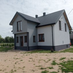 Usługi Remontowo-Wykończeniowe Damian Zaborowski - Dwukondygnacyjny dom z szarą i białą elewacją, ciemną podmurówką i brązowymi ramami okiennymi, otoczony zielenią i piaszczystym terenem.
