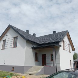 Usługi Remontowo-Wykończeniowe Damian Zaborowski - Nowoczesny dom jednorodzinny z szarą elewacją i ciemną dachówką w Grudusku. Widoczne detale wykończenia, schody wejściowe i fragment samochodu na pierwszym planie.
