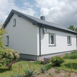 Usługi Remontowo-Wykończeniowe Damian Zaborowski - Biały dom jednorodzinny z szarym dachem i satelitą na elewacji, otoczony zielenią. Widok z boku, w słoneczny dzień. Świeżo pomalowana elewacja.