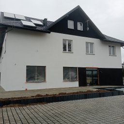 Usługi Remontowo-Wykończeniowe Damian Zaborowski - Dwukondygnacyjny budynek z białą elewacją i czarnym dachem, panele słoneczne na dachu, okna zabezpieczone folią, kostka brukowa przed budynkiem.
