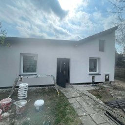 Usługi Remontowo-Wykończeniowe Damian Zaborowski - Dom jednorodzinny po malowaniu elewacji, widoczne drabiny, wiaderka z farbą i świeżo pomalowana fasada w odcieniach bieli i szarości. Ujęcie z poziomu gruntu.