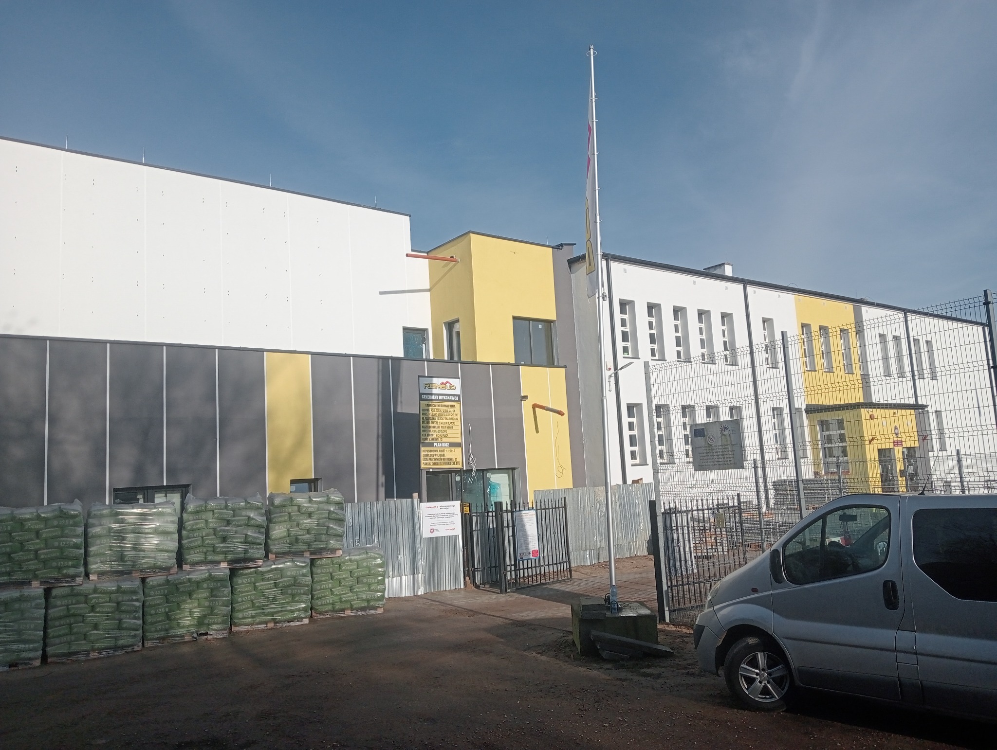 Budynek po renowacji z elementami ocieplenia, widoczne worki z materiałami budowlanymi i zaparkowany samochód dostawczy. Tablica informacyjna na elewacji.