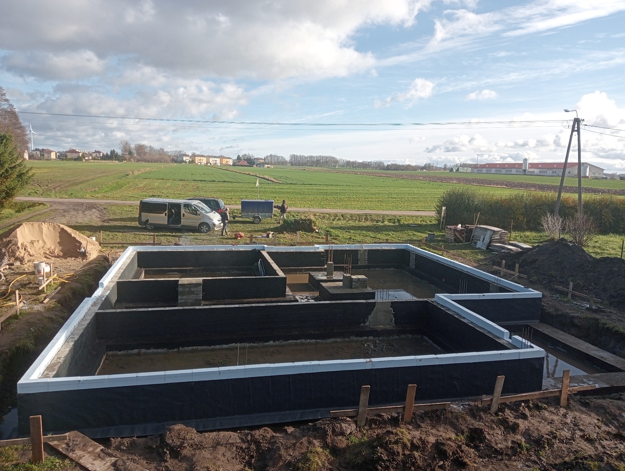 Fundamenty domu jednorodzinnego z izolacją przeciwwilgociową, widok z góry na tle krajobrazu wiejskiego z polem i zabudowaniami. Etap prac budowlanych.