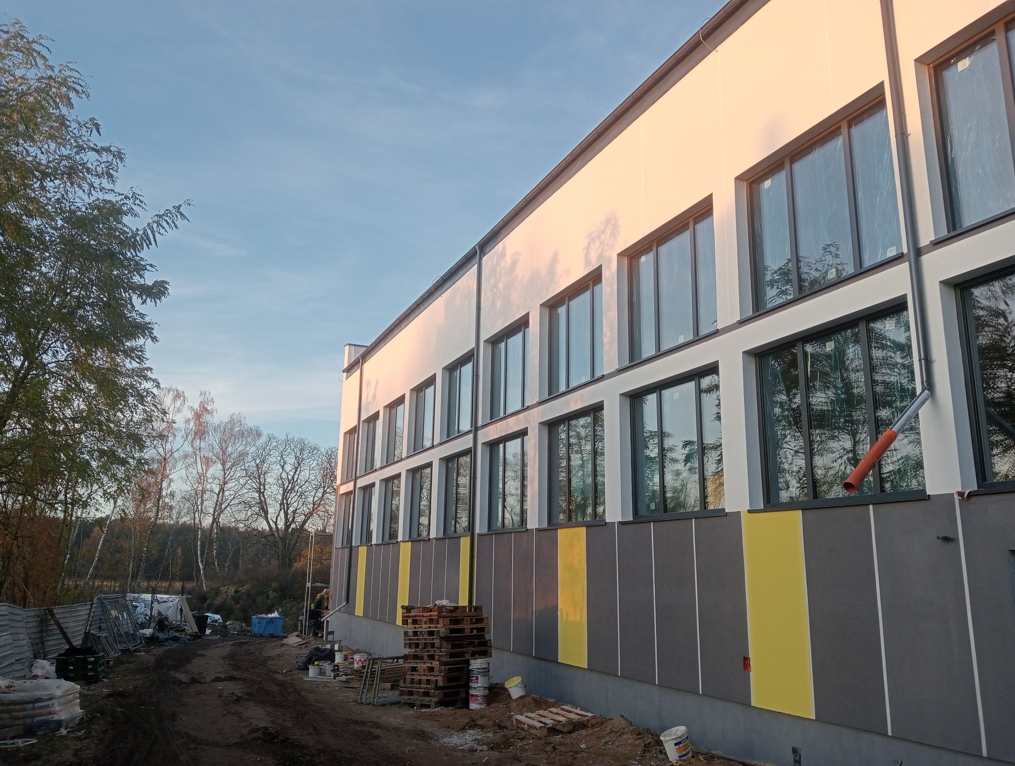 Nowoczesny budynek z oknami i elewacją w odcieniach szarości i żółci, w trakcie prac wykończeniowych. Widoczne palety i materiały budowlane. Ujęcie z dołu, w tle drzewa.