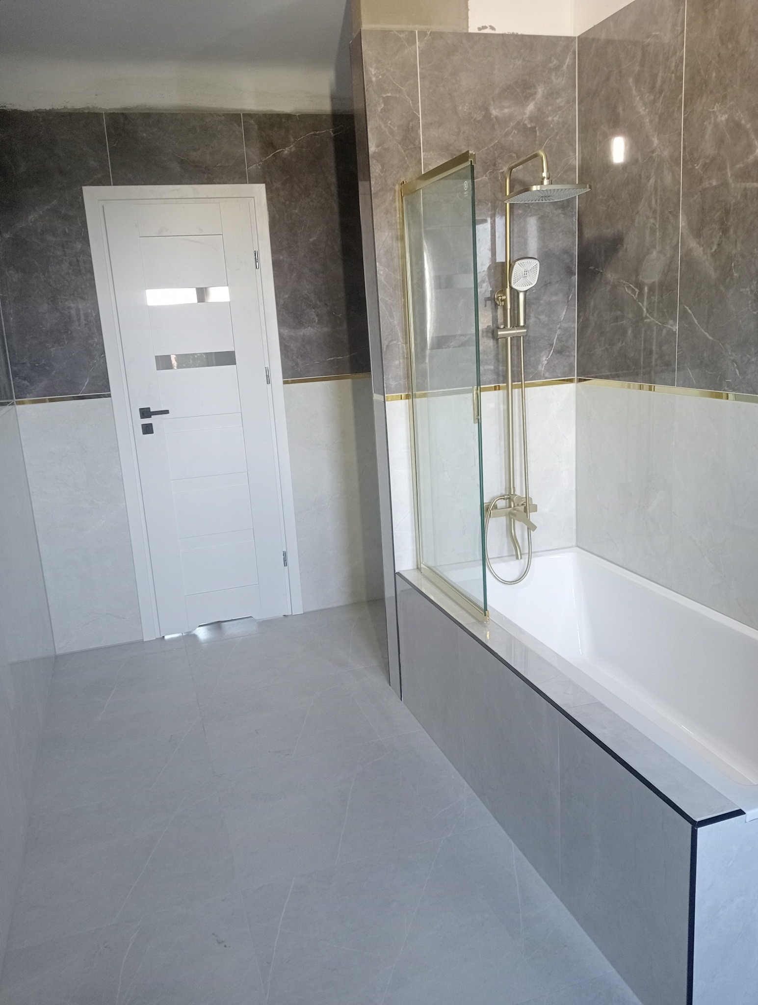 Wykończona łazienka z wanną obudowaną szarą płytką, złotą armaturą i szklanym parawanem. Na ścianach szare i białe płytki imitujące marmur, białe drzwi z przeszkleniami.