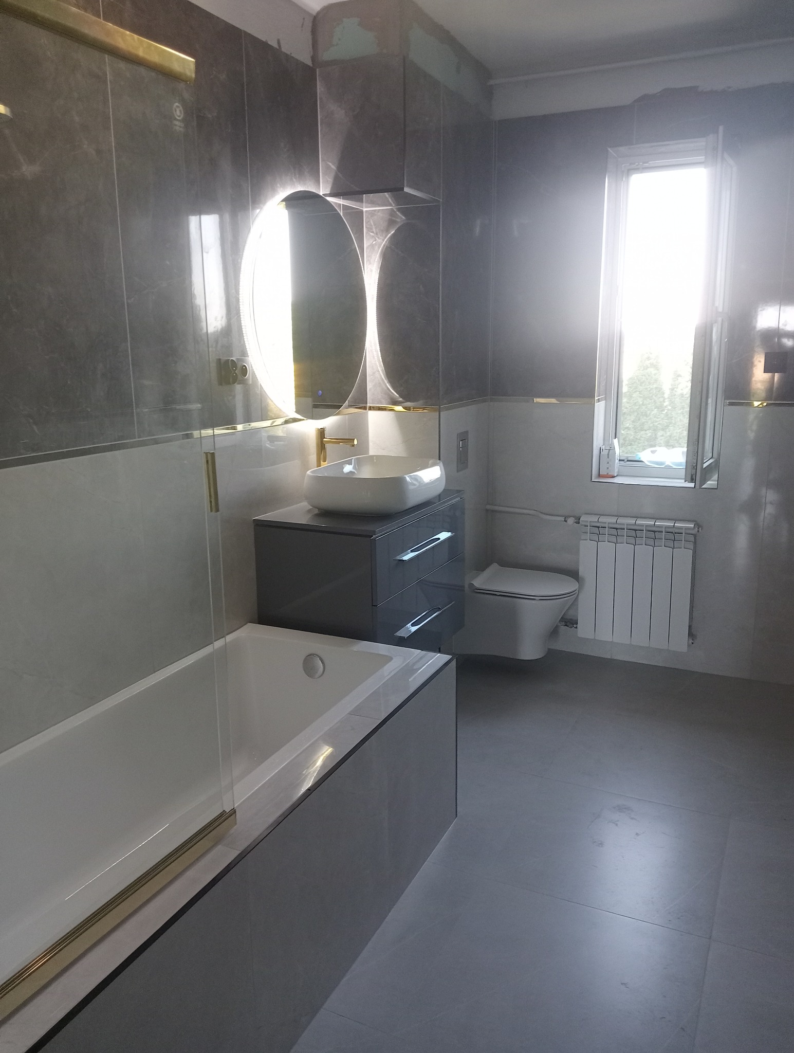 Nowoczesna łazienka w szarościach z podświetlanym lustrem, białą umywalką nablatową i grzejnikiem. Widoczne płytki imitujące marmur i złote akcenty.