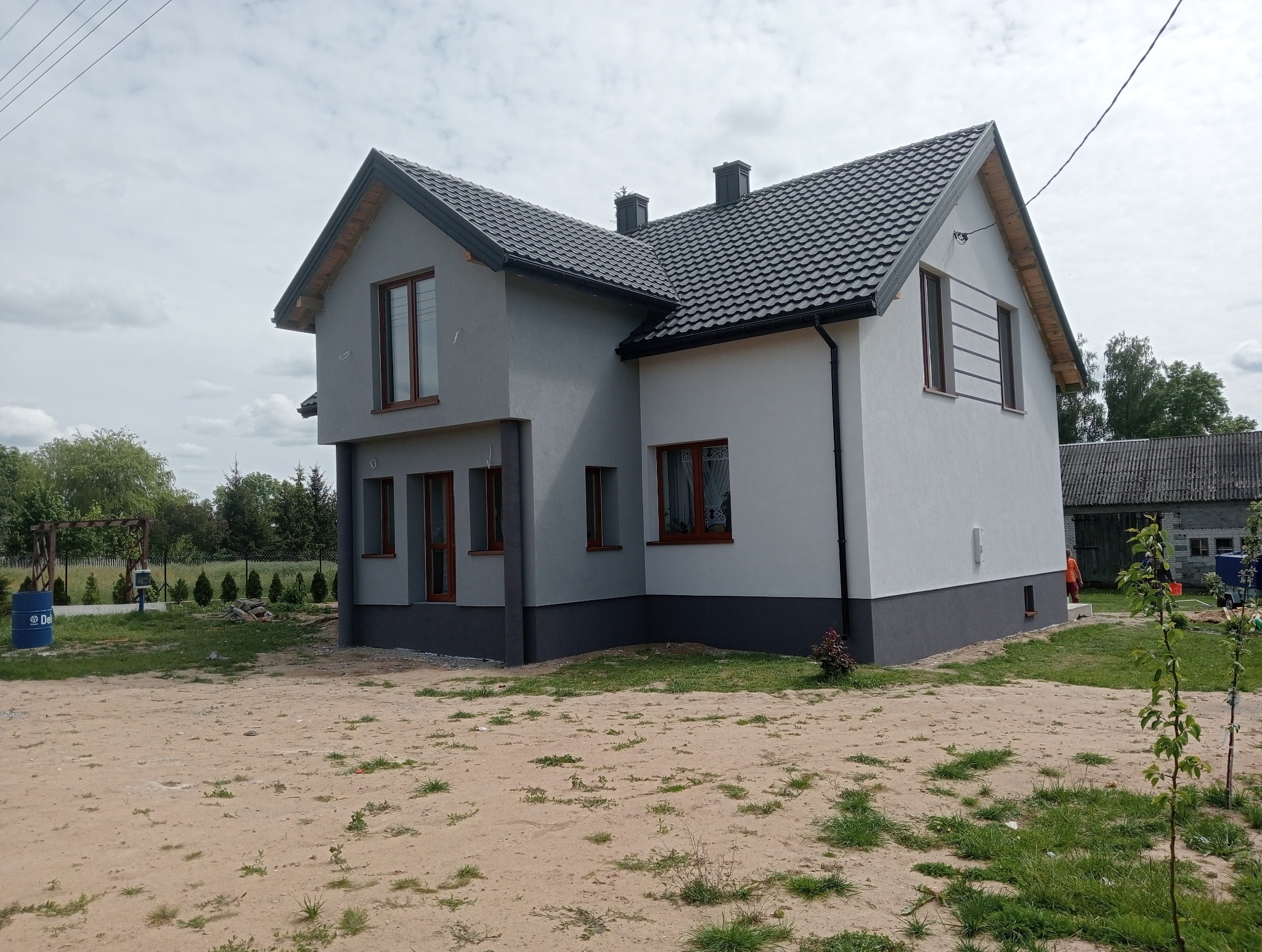 Dwukondygnacyjny dom z szarą i białą elewacją, ciemną podmurówką i brązowymi ramami okiennymi, otoczony zielenią i piaszczystym terenem.