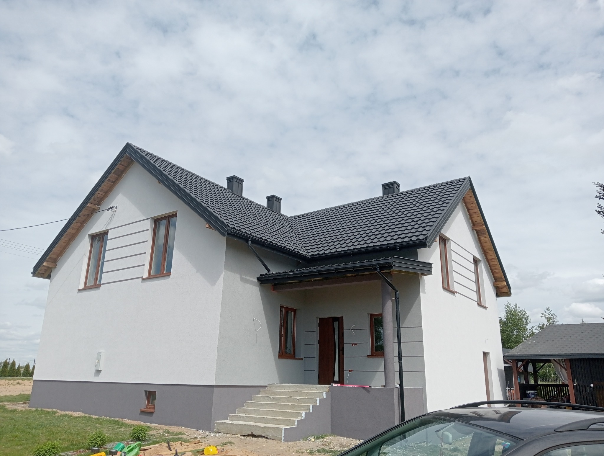 Nowoczesny dom jednorodzinny z szarą elewacją i ciemną dachówką w Grudusku. Widoczne detale wykończenia, schody wejściowe i fragment samochodu na pierwszym planie.