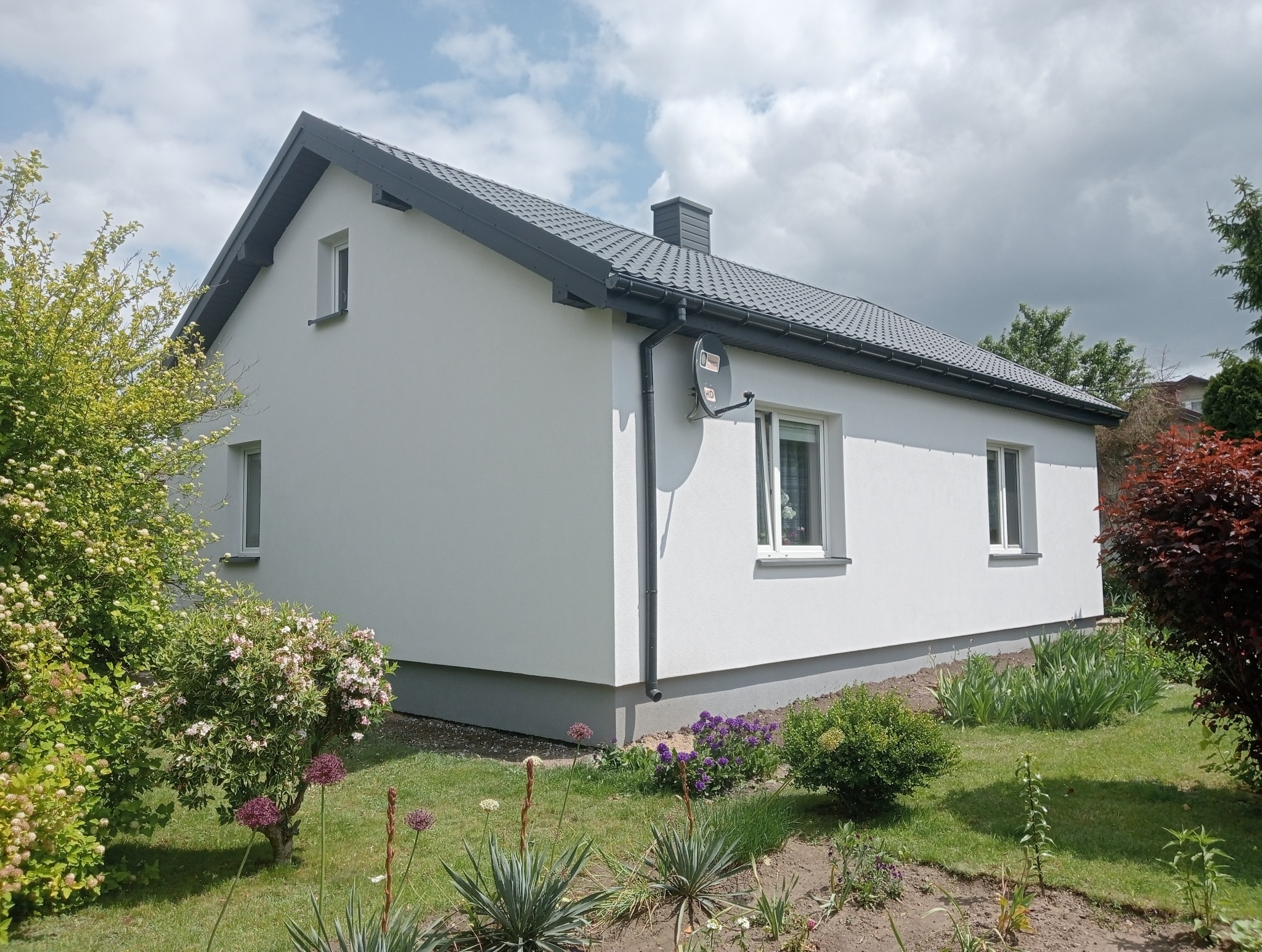 Biały dom jednorodzinny z szarym dachem i satelitą na elewacji, otoczony zielenią. Widok z boku, w słoneczny dzień. Świeżo pomalowana elewacja.