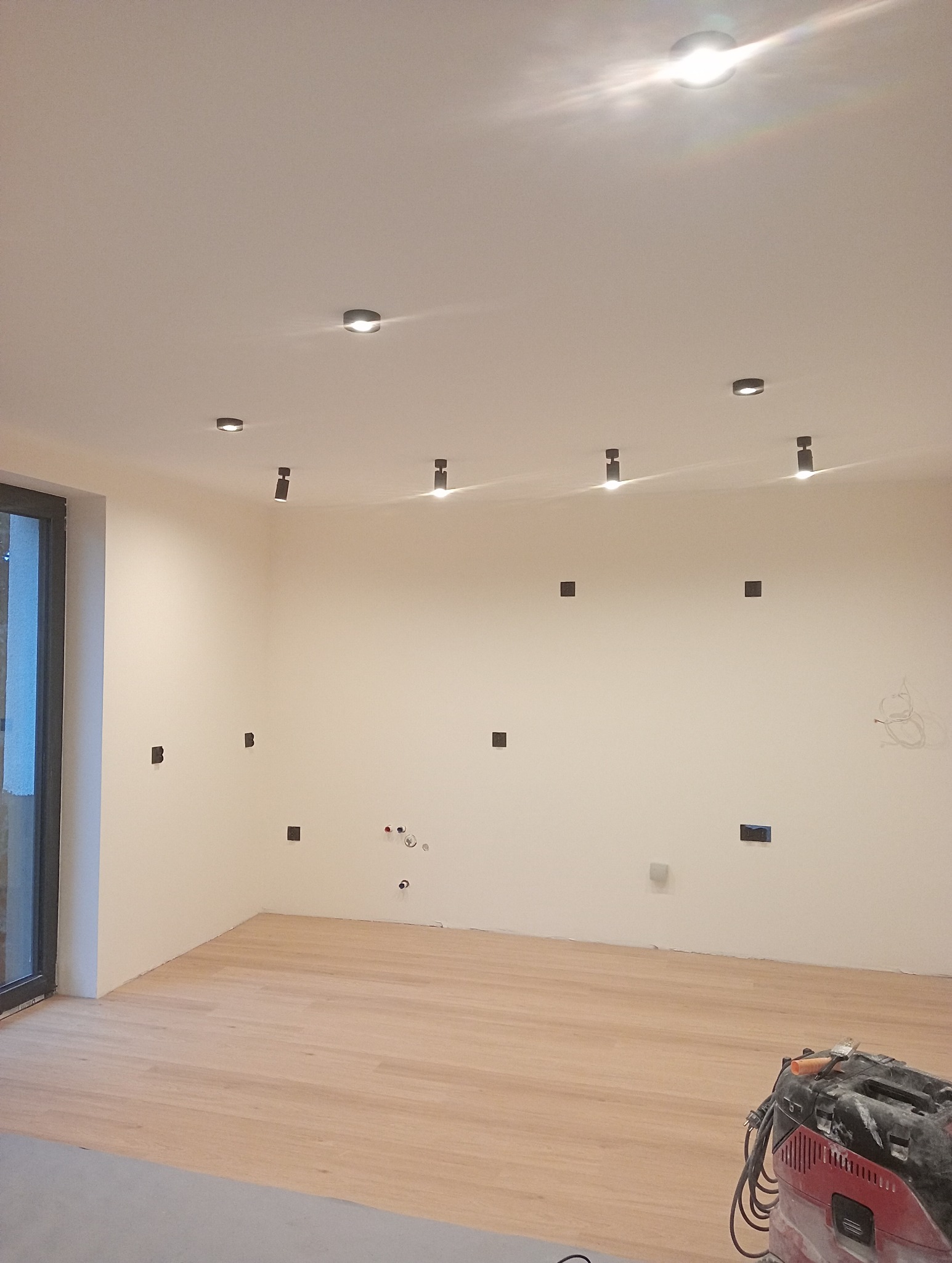 Jasne panele podłogowe w nowym wnętrzu z zamontowanym oświetleniem punktowym. Widoczne puszki elektryczne i odkurzacz przemysłowy w rogu pomieszczenia.