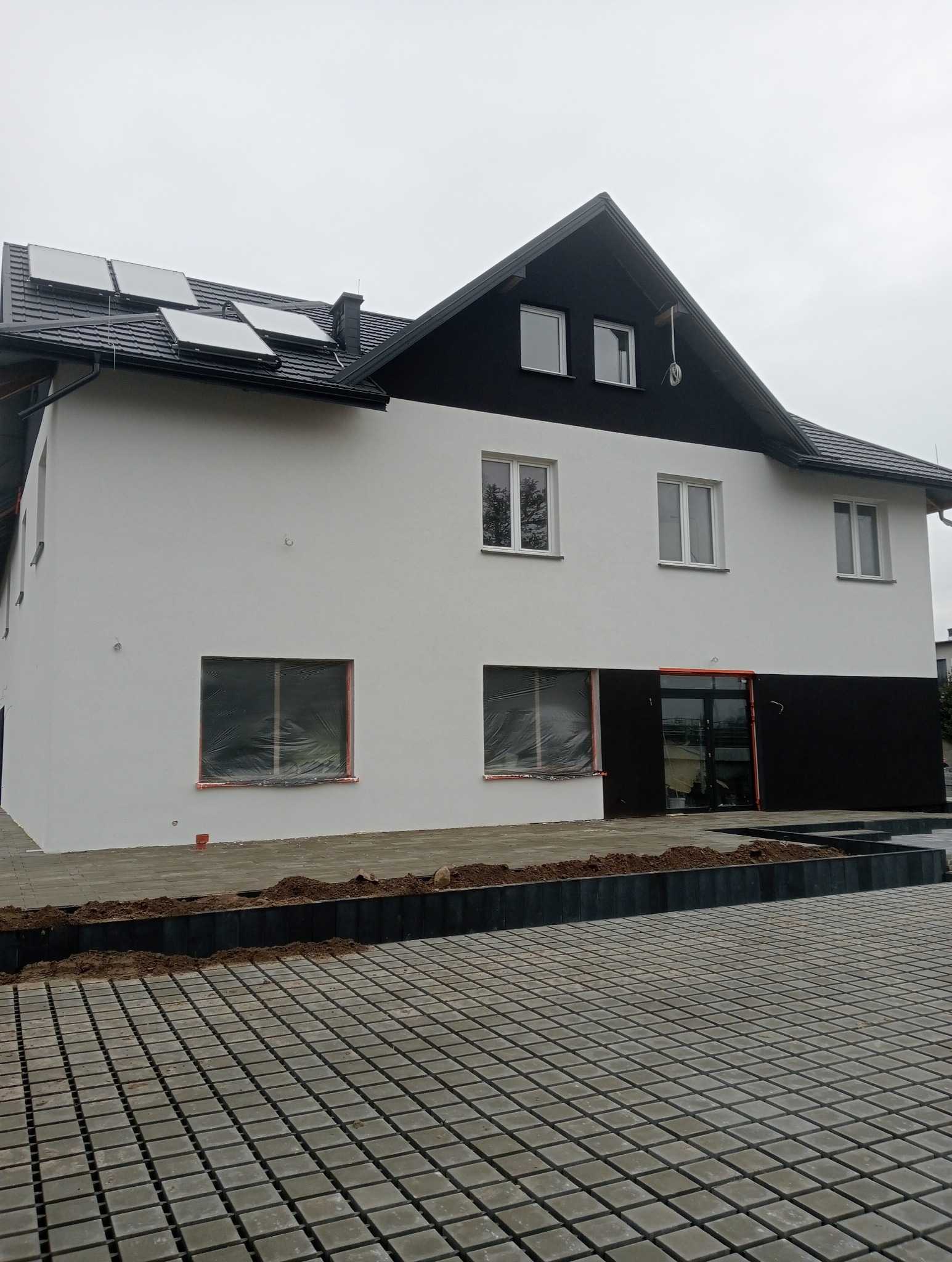 Dwukondygnacyjny budynek z białą elewacją i czarnym dachem, panele słoneczne na dachu, okna zabezpieczone folią, kostka brukowa przed budynkiem.